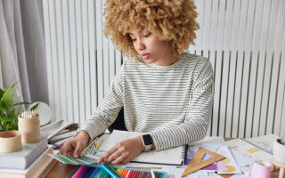 Creative female designer works on interior project chooses materials and details works with colour palette has serious expression poses at desktop against cozy interior. Renovation and decor concept
